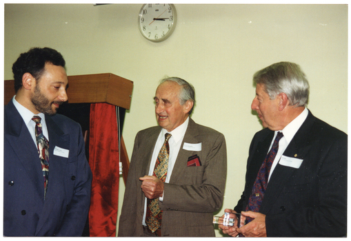 COL photo of three men standing near a commemorative plaque on a display board. One of the men is holding a small clear glass or perspex trophy in his hand. There is a clock on the wall behind them, with the hand positioned at 3:15.