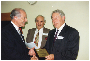 COL photo of two men shaking hands while another man, standing between them, looks on. One of the men is holding a small brass plaque in his other hand. On the wall behind them is a black display board with a wooden pelmet at the top and on the wall above them is a clock (both partially obscured).