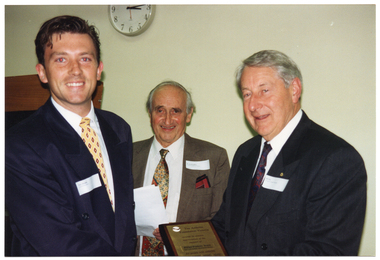 COL photo of three men standing close together. One of the men is holding a small brass plaque in his hand while another man is holding some sheets of paper in his hands. On the wall behind them is a black display board with a wooden pelmet at the top and on the wall above them is a clock (both partially obscured).