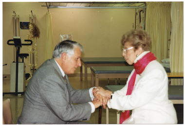 COL photo of a man and a woman, sitting down. They  are clasping both of their hands to one another's. They are in a hospital ward. Behind them are three hospital examination tables with booth curtains, and an electronic exercise bike. Hanging from a pole behind the bike is a plastic skeleton with a curly brown wig on its head.