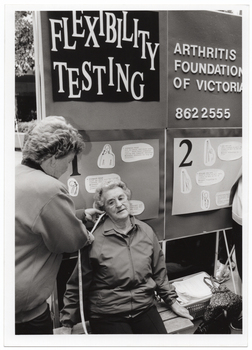 B&W photo of a woman using a measuring tape to measure the distance between another woman's ear and shoulder. Behind them is a display board with the words "Flexibility testing", "Arthritis Foundation of Victoria", the telephone number "862 2555", and various sketch drawings with corresponding text.