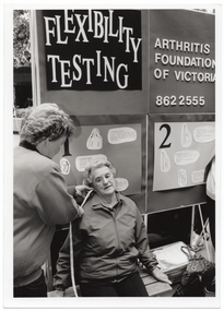 B&W photo of a woman using a measuring tape to measure the distance between another woman's ear and shoulder. Behind them is a display board with the words "Flexibility testing", "Arthritis Foundation of Victoria", the telephone number "862 2555", and various sketch drawings with corresponding text.