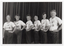 B&W photo of a small group of people posing for a photo. They are standing in a row in three-quarter profile, each with one hand on their left hip. Behind them is a large curtain, similar to a stage curtain.