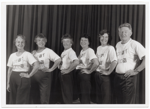 B&W photo of a small group of people posing for a photo. They are standing in a row in three-quarter profile, each with one hand on their left hip. Behind them is a large curtain, similar to a stage curtain.