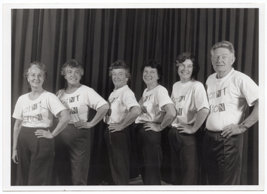 B&W photo of a small group of people posing for a photo. They are standing in a row in three-quarter profile, each with one hand on their left hip. Behind them is a large curtain, similar to a stage curtain.