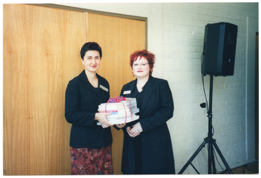 COL photo of two women, standing. One of them is holding a stack of books with some purple and pink ribbon wrapped around it. Behind them, there is a black speaker on a vertical stand. In the background, there is a large wooden door and a white-painted brick wall.