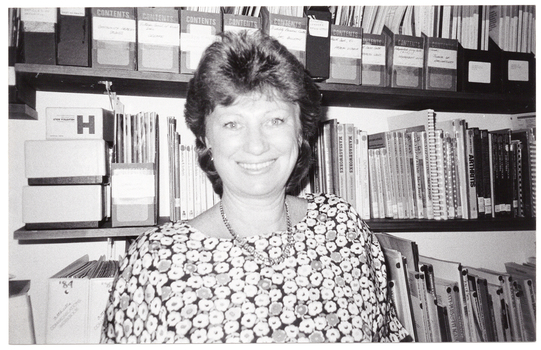 B&W photo of a woman standing in front of several wall shelves containing books and other print resources.