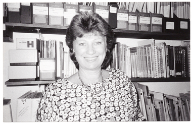 B&W photo of a woman standing in front of several wall shelves containing books and other print resources.