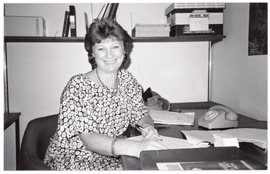 B&W photo of a woman sitting at a desk on which there is a telephone and some paperwork. She is holding a pen, and facing the camera. Behind her is a high shelf containing various folders, telephone directories, and an archive box.