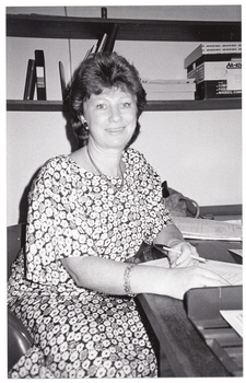 B&W photo of a woman sitting at a desk, on which is some paperwork. She is holding a pen, and facing the camera. On the wall behind her are two high shelves containing various folders, telephone directories, and an archive box.