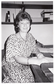 B&W photo of a woman sitting at a desk, on which is some paperwork. She is holding a pen, and facing the camera. On the wall behind her are two high shelves containing various folders, telephone directories, and an archive box.