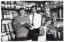 B&W photo of two women and a man, with some books. They are standing behind a table, in front of two walls of shelves full of books, journals, and other printed materials. The man and one of the women are holding two copies of the Collier's Dictionary. The other woman has her hands on an upright copy of the New Illustrated Everyman's Encyclopedia, which is on the table. In front of the encyclopedia, there is a stack of books on the table, and another short stack of two books beside it.