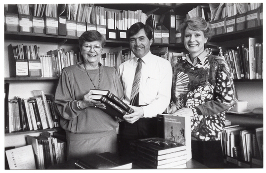 B&W photo of two women and a man, with some books. They are standing behind a table, in front of two walls of shelves full of books, journals, and other printed materials. The man and one of the women are holding two copies of the Collier's Dictionary. The other woman has her hands on an upright copy of the New Illustrated Everyman's Encyclopedia, which is on the table. In front of the encyclopedia, there is a stack of books on the table, and another short stack of two books beside it.
