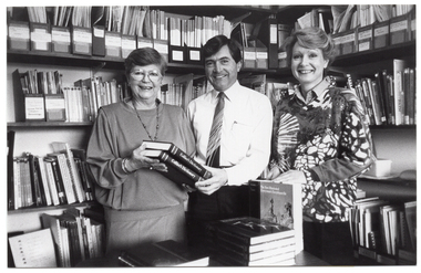 B&W photo of two women and a man, with some books. They are standing behind a table, in front of two walls of shelves full of books, journals, and other printed materials. The man and one of the women are holding two copies of the Collier's Dictionary. The other woman has her hands on an upright copy of the New Illustrated Everyman's Encyclopedia, which is on the table. In front of the encyclopedia, there is a stack of books on the table, and another short stack of two books beside it.