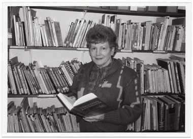 B&W photo of a woman holding an open book, which is titled "Treating Arthritis". She is standing in front of some rows of shelving filled with books.