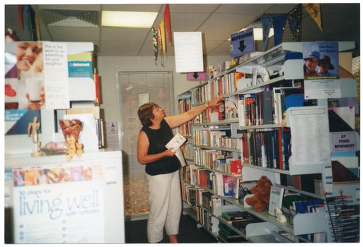 COL photo of a woman reaching for some books on the top shelf of a row of shelving. The shelves are filled with books and other printed resources. There is a brown teddy bear on one of the shelves. On the end of the rows of shelving, as well as around the room and hanging from the ceiling, there are various posters, signs, and some bunting.