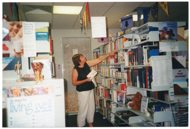 COL photo of a woman reaching for some books on the top shelf of a row of shelving. The shelves are filled with books and other printed resources. There is a brown teddy bear on one of the shelves. On the end of the rows of shelving, as well as around the room and hanging from the ceiling, there are various posters, signs, and some bunting.