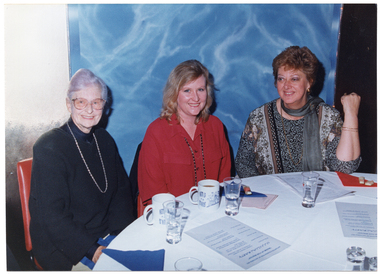 COL photo of three women sitting at a round table covered with a white tablecloth. On the table, there are some printed sheets of paper, mugs of tea or coffee, glasses of water, and some finger food on top of paper serviettes.