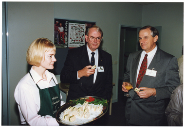 COL photo of three people standing in a room.  A woman, wearing a dark green 'Arthritis Foundation of Victoria' apron, is passing around a tray of sandwiches. Facing her  are two men, each with a sandwich in one hand. One of the men is also holding a wine glass filled with orange juice in his other hand. The men are both wearing suits with a name label affixed to the front. There are other people also in the room (partially obscured), to the right of the frame.