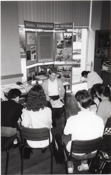 B&W photo of a small group of people seated in a semi-circle. Facing them is a woman, also seated. Behind her, there is a table with various printed materials on it. Behind the table, there is a large freestanding Arthritis Foundation of Victoria display board. There are Photographs and posters on the display board, and a sign with the text, "WATER EXERCISE". Behind the display board are tall floor to ceiling windows, with vertical blinds covering the top windows.