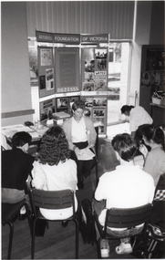 B&W photo of a small group of people seated in a semi-circle. Facing them is a woman, also seated. Behind her, there is a table with various printed materials on it. Behind the table, there is a large freestanding Arthritis Foundation of Victoria display board. There are Photographs and posters on the display board, and a sign with the text, "WATER EXERCISE". Behind the display board are tall floor to ceiling windows, with vertical blinds covering the top windows.