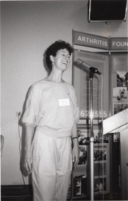 B&W photo of a woman speaking into a microphone on a stand. Behind her is an Arthritis Foundation of Victoria display board with Photographs and posters.