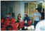 COL photo of some several people in an almost empty room, with several rows of chairs. A woman, who is seated, is twisting around to speak to a man who is sitting in the row behind her. Another woman, who is sitting in the row in front of her, is looking down to read a publication. There are some big windows and an open doorway leading to a courtyard. A woman is walking into the room from the courtyard, while another woman is standing in the doorway.