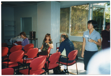 COL photo of some several people in an almost empty room, with several rows of chairs. A woman, who is seated, is twisting around to speak to a man who is sitting in the row behind her. Another woman, who is sitting in the row in front of her, is looking down to read a publication. There are some big windows and an open doorway leading to a courtyard. A woman is walking into the room from the courtyard, while another woman is standing in the doorway.