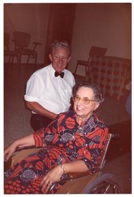 COL photo of a man and a woman smiling broadly. He is wearing a white shirt with a black bow tie and black trousers, while she is wearing a colourful red and blue dress. She is a wheelchair user, and he appears to be kneeling beside her. In the background are some chairs and a table covered in a colourful tablecloth.