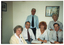 COL photo of a small group of people sit or stand around a small table, on which there are some printed papers.