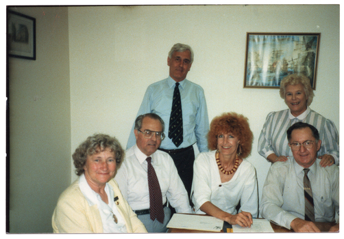 COL photo of a small group of people sit or stand around a small table, on which there are some printed papers.