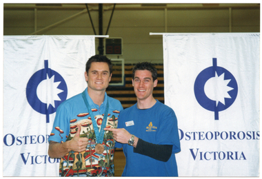 COL photo of two men standing in front of two large, vertical, freestanding banners. They are both smiling broadly. The man to the left of the frame is wearing a colourful shirt with a large bronze Olympic medal on a blue ribbon hanging from his neck. He is doing a "thumbs up" action with his hand. The man on the right of the frame is wearing a blue Arthritis Victoria t-shirt. He is pointing to the other man. Both banners display the text, "Osteoporosis Victoria", with a corresponding logo. There is stadium seating in the background.