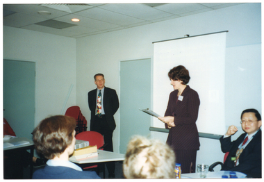 COL photo of a man and a woman standing in a large room, facing a group of people who are seated. Behind the woman, there is a projector screen on a stand, and there is a stack of chairs in the corner. In front of the woman, there is a table with a stack of books on it. In the foreground, a few people (partially obscured) have been Photographed from behind.