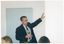 COL photo of a man holding one arm up at a 45 degree angle. He is holding a remote control in his other hand. In the foreground, a few people (partially obscured) have been Photographed from behind.
