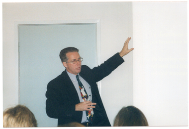 COL photo of a man holding one arm up at a 45 degree angle. He is holding a remote control in his other hand. In the foreground, a few people (partially obscured) have been Photographed from behind.