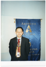 COL photo of a man, smiling, standing in front of a bright blue and gold Arthritis Victoria/Osteoporosis Victoria banner. He is wearing a lanyard with a name with his photo on it.