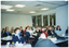 COL photo of a group of people inside a large room. They are seated at tables, on which there are various items, including drinks, napkins, and paperwork.  There is a large rectangular window along one wall, near the back corner of the room.