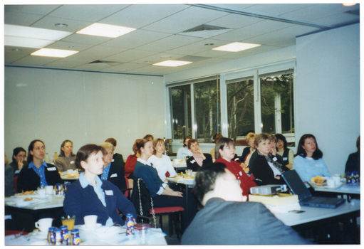 COL photo of a group of people inside a large room. They are seated at tables, on which there are various items, including drinks, napkins, and paperwork.  There is a large rectangular window along one wall, near the back corner of the room.