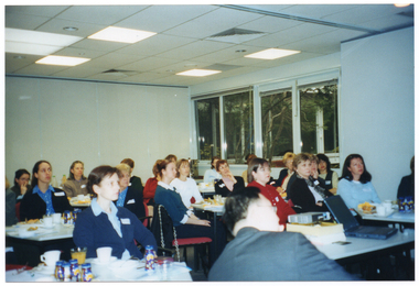 COL photo of a group of people inside a large room. They are seated at tables, on which there are various items, including drinks, napkins, and paperwork.  There is a large rectangular window along one wall, near the back corner of the room.