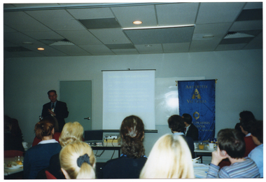 COL photo of a man making a presentation to a roomful of seated people. There is a screen beside him, onto which some text is been projected from a slide projector on a table. Beside the screen, there is a freestanding Arthritis Victoria/Osteoporosis Victoria blue and gold banner.