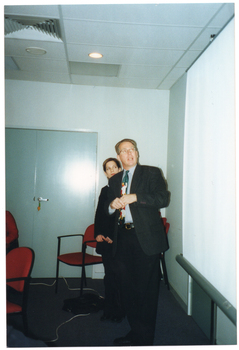 COL photo of a man and a woman (partially obscured) looking up at a slide projector screen. The man is standing in front of the woman. There are some chairs and a set of closed double doors behind them. There are some more chairs in the foreground, to the left of the frame.