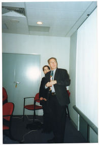 COL photo of a man and a woman (partially obscured) looking up at a slide projector screen. The man is standing in front of the woman. There are some chairs and a set of closed double doors behind them. There are some more chairs in the foreground, to the left of the frame.