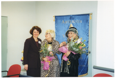 COL photo of three women posing for a photo in front of a large blue and gold freestanding vertical banner. Two of the women are each holding a bouquet of flowers. On the banner, there is the text "ARTHRITIS", with the remaining text obscured.