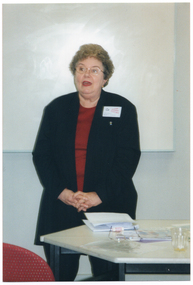 COL photo of a woman, standing in front of a projector screen, speaking. She is wearing a name badge with the text, "MELBOURNE OSTEOPOROSIS SUPPORT GROUP" and "PRESIDENT, BERYL LOGIE".