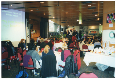 COL photo of a large group of people sitting at several large round tables, while others are standing in the background. On the left, there is a large screen, with a slide being projected onto it. It contains the text, "Osteoporosis Victoria and MONSU present Strong Bones Beautiful Body Breakfast', as well as the Arthritis Victoria and Osteoporosis Victoria logos. To the right of the screen, there is a vertical, freestanding banner with the Osteoporosis Victoria logo and a few balloons. In the background on the right, there is a black and yellow banner hanging from a desk or counter with the text 'MONASH UNIVERSITY'. In the foreground, to the right of the frame, there is a table containing a range of labelled fresh and packaged food items. Behind the table, there is a display board with a large Osteoporosis Victoria logo and some smaller printed sheets of paper.