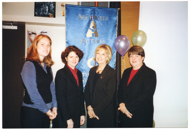 COL photo of four women posing for a photo in front of a blue and gold Arthritis Victoria/Osteoporosis Victoria banner. There are some balloons next to the banner.