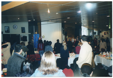 COL photo of a large group of people standing around several large tables. On top of the tables, there are cups and sauces and other miscellaneous items. One table to the right of the frame contains a display of labelled fresh and packaged food items. In the background, there is a woman standing near a large freestanding blue and gold banner, a few balloons, and a speaker  on a tall stand.
