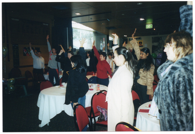 COL photo of a large group of people standing around several large tables. They each have one arm raised up towards the ceiling. On top of the tables, there are cups and sauces and other miscellaneous items. There are some balloons in the background, and an exit sign affixed to the ceiling or up high on a wall.