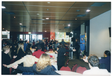 COL photo of a large group of people standing around several large tables. One woman is standing to the right of the frame, near a large freestanding blue and gold banner, some balloons, and a speaker on a tall stand. They all have their arms outstretched to the side. On top of the tables, there are cups and sauces and other miscellaneous items. In the background, there are several large windows along one wall.