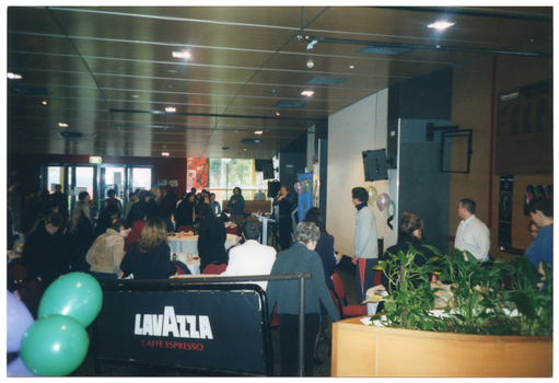 COL photo of a large group of people standing around several large tables. One woman is standing in the centre of the frame,  near a large freestanding blue and gold banner, some balloons, and a speaker on a tall stand. On top of the tables, there are cups and sauces and other miscellaneous items. In the background, there are several large windows along one wall. In the foreground, there are some balloons to the left of the frame, a Lavazzo branded barrier/room divider in the centre of the frame, and a large circular planter containing several potted plants to the right of the frame.